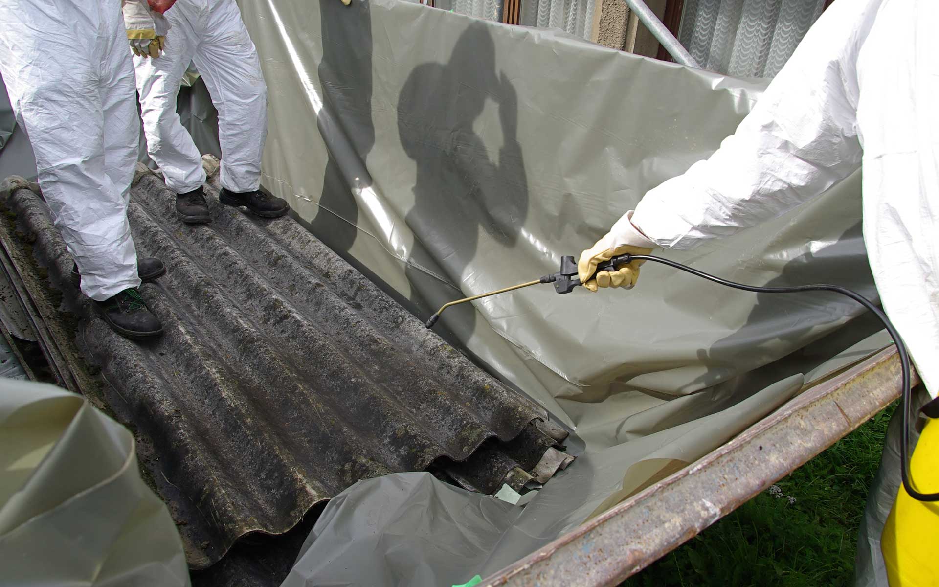 Das Foto für den Asbest Auffrischungskurs zeigt Arbeiter in weißen Schutzanzügen, die Chemikalien auf Beton sprühen.